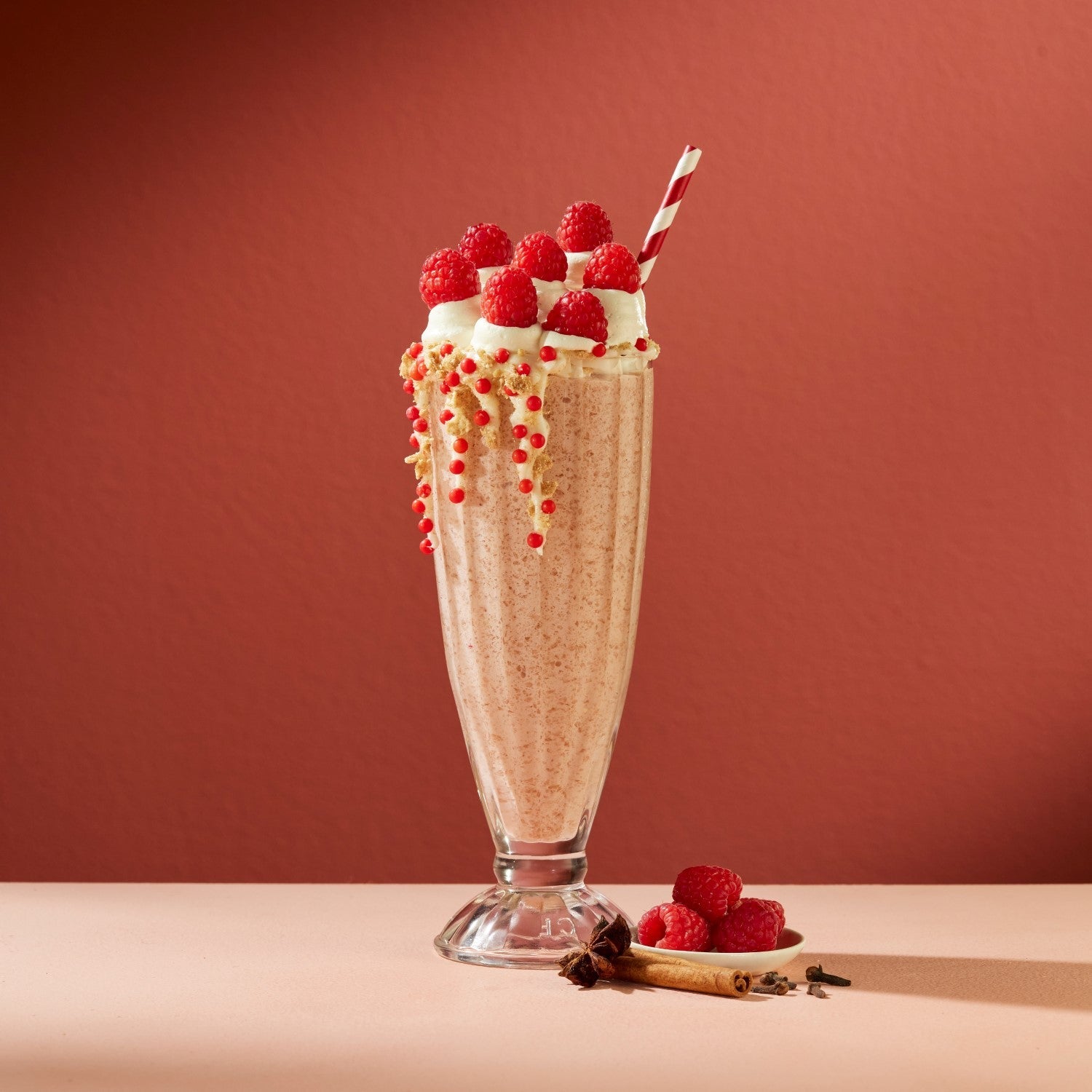 A light pink raspberry chai shake in a classic milkshake glass topped with fresh raspberries standing out in front of a solid red background